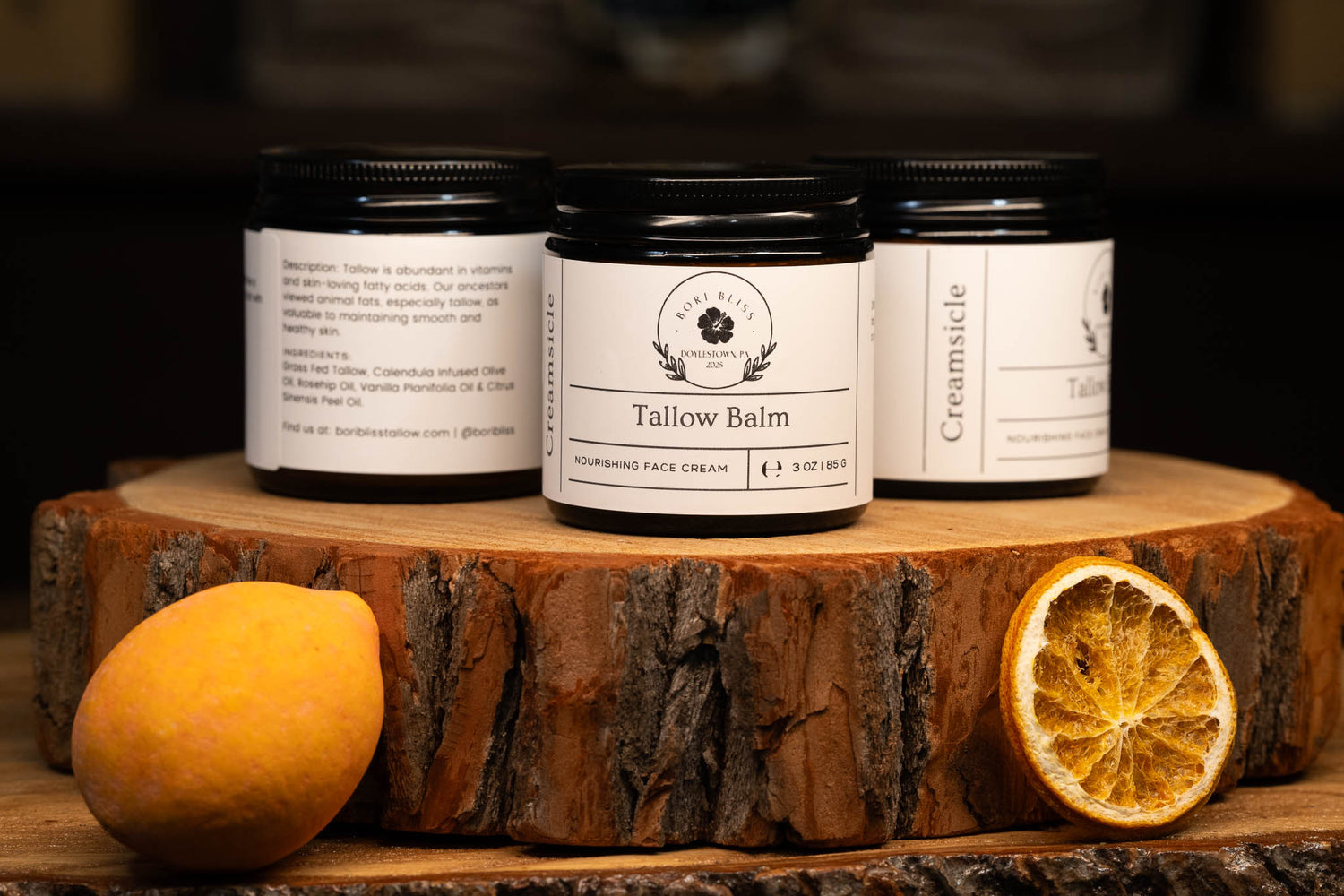 Three 3oz jars of Bori Bliss Creamsicle tallow balm on a wooden stand with oranges on a dark background